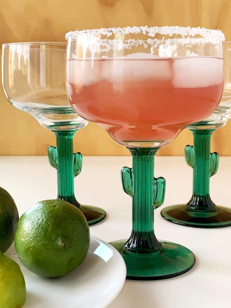 A pink margarita on the rocks made in a cactus margarita glass, in front of two other margarita glasses, next to some limes