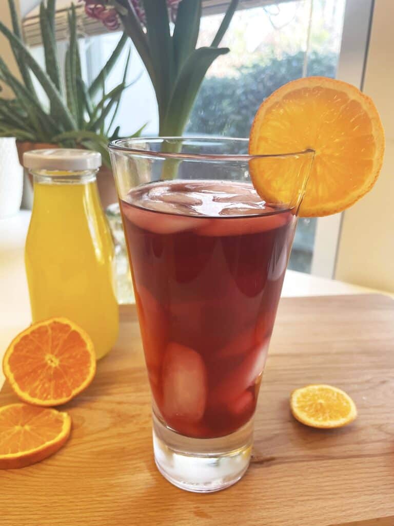 Glass of Iced Mandarin Hibiscus Tea garnished with an orange slice, with a bottle of homemade mandarin syrup in the background