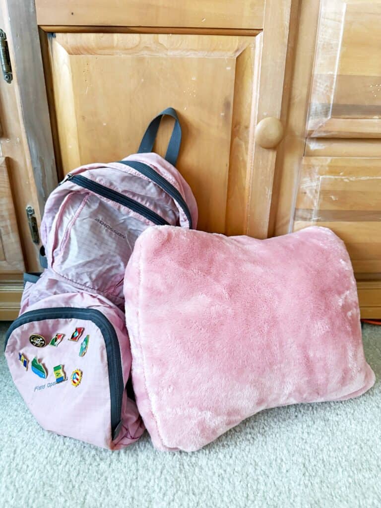 A pink travel blanket that folds up into a pillow, next to a pink waterproof backpack